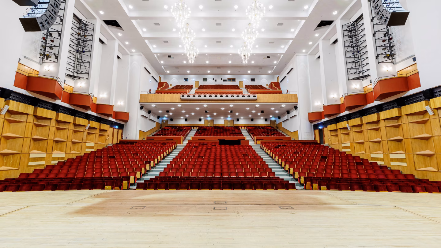 A brightly-lit auditorium with red seats, two banks of stalls seating and a balcony.
